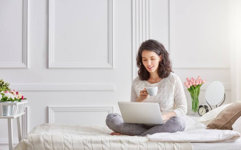 Woman drinking a cup of coffee while sitting in a bed, using her computer: 7 stressful blogging mistakes new bloggers make. 